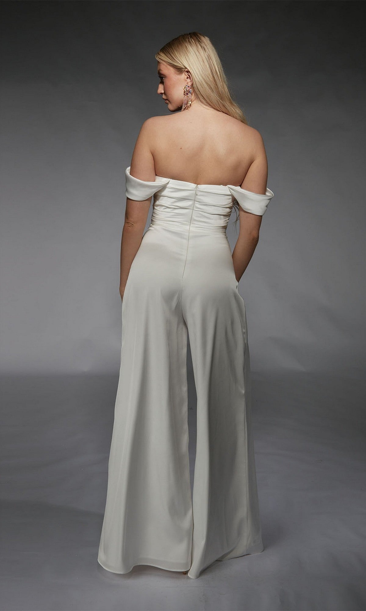 A woman faces away, wearing the white Long Formal Jumpsuit: Alyce 70071 with off-shoulder design and wide-leg pants against a plain dark background.